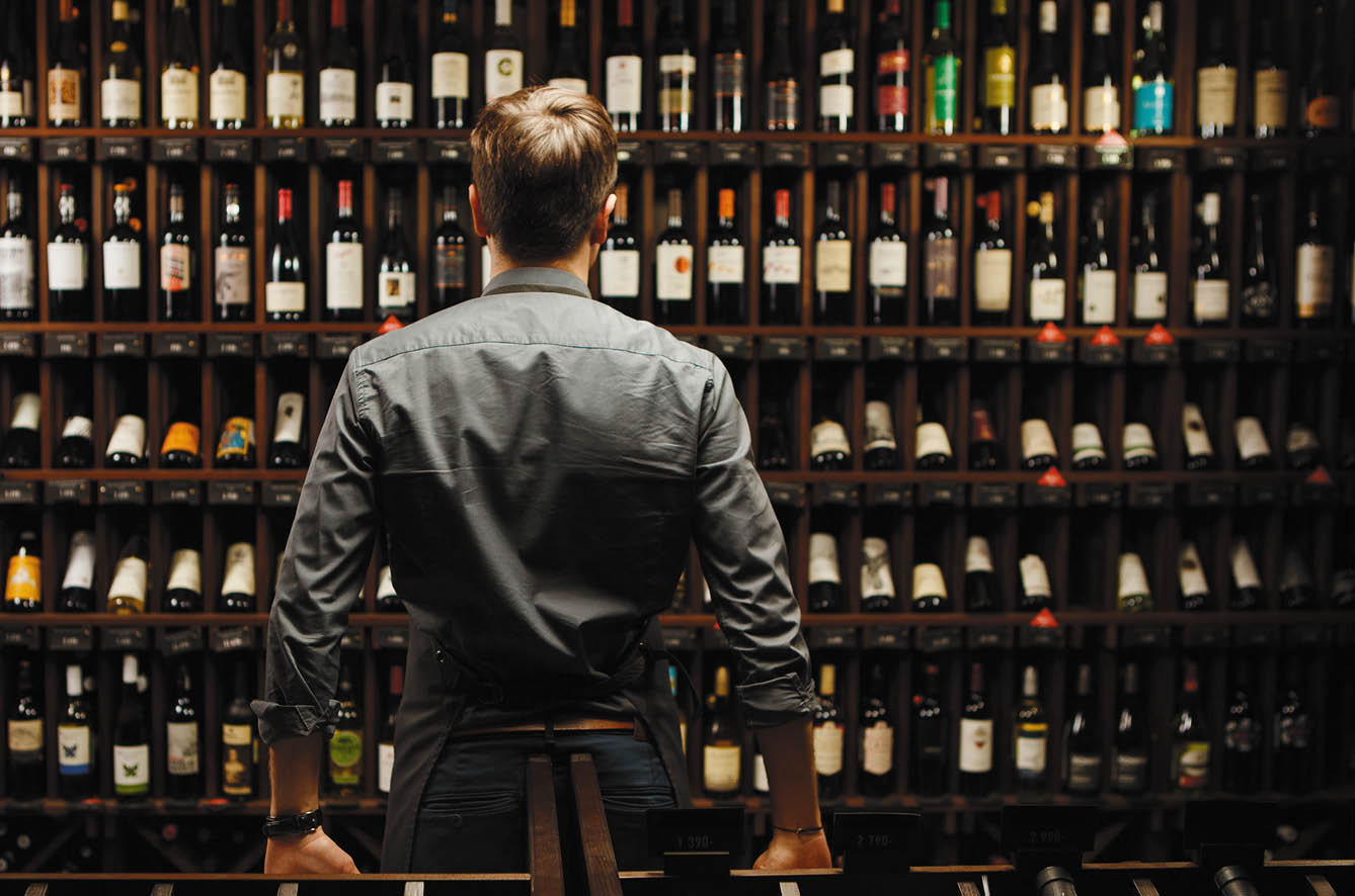 Bartender at wine cellar full of bottles with exquisite alcohol drinks that have various sweet and sour tastes and dates of manufacture on large wooden shelves 