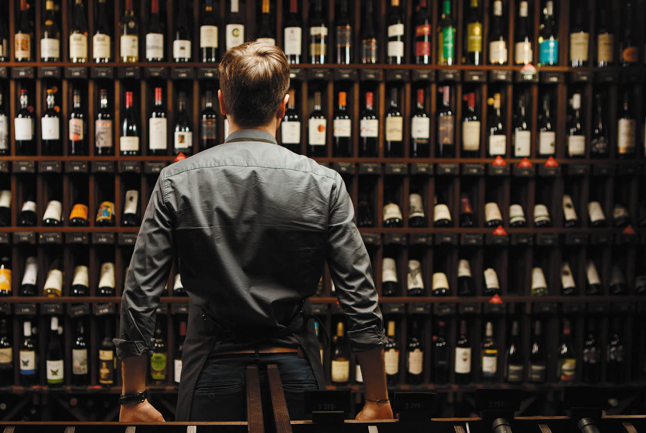Bartender at wine cellar full of bottles with exquisite alcohol drinks that have various sweet and sour tastes and dates of manufacture on large wooden shelves 
